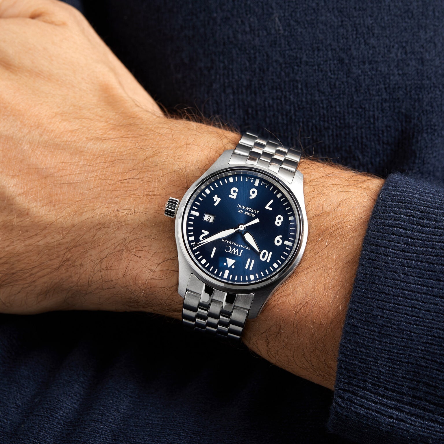 Someone wearing the IWC Schaffhausen Pilots Watch Mark XX—a silver aviation watch with a blue dial, white numerals, and metal link band—paired with a dark blue long-sleeve shirt.