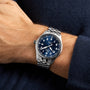 Someone wearing the IWC Schaffhausen Pilots Watch Mark XX—a silver aviation watch with a blue dial, white numerals, and metal link band—paired with a dark blue long-sleeve shirt.