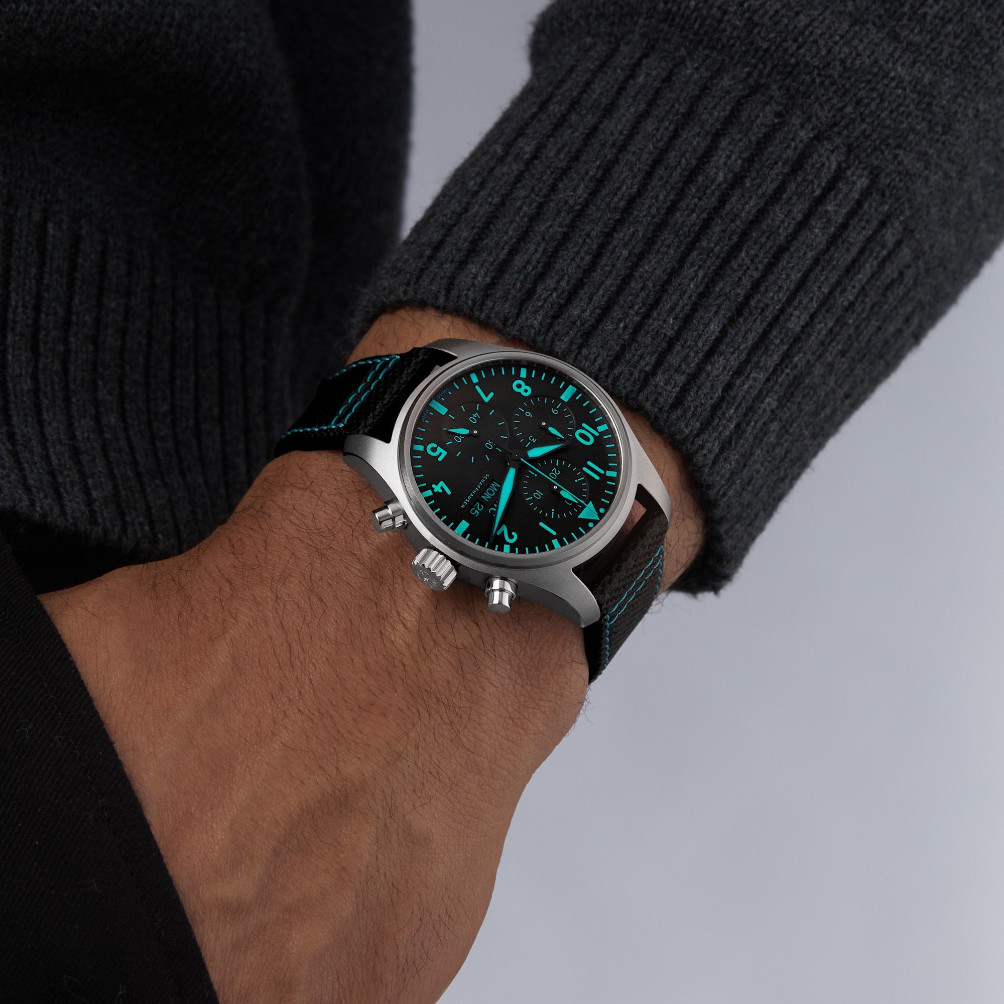 A person in a dark sweater showcases the IWC Schaffhausen Pilots Watch Chronograph 41 Edition Mercedes - AMG PETRONAS Formula One™ Team with a black strap and luminous blue-green dial displaying 10:09.