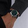 A person in a dark sweater showcases the IWC Schaffhausen Pilots Watch Chronograph 41 Edition Mercedes - AMG PETRONAS Formula One™ Team with a black strap and luminous blue-green dial displaying 10:09.