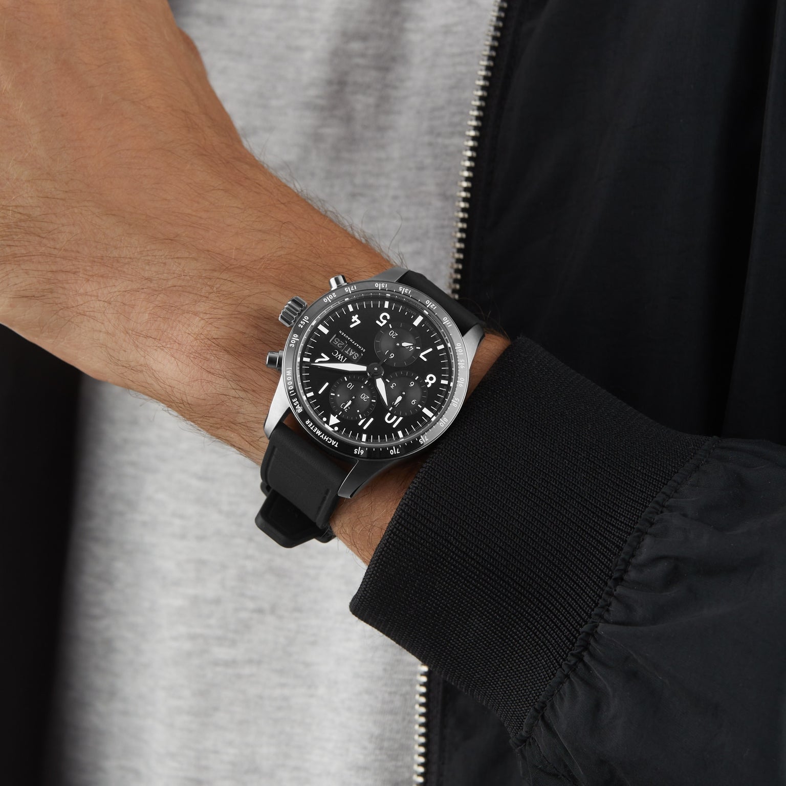A person wearing an IWC Schaffhausen Pilots Watch Performance Chronograph 41 AMG with a black strap and sapphire glass, paired with a black jacket over a grey shirt.