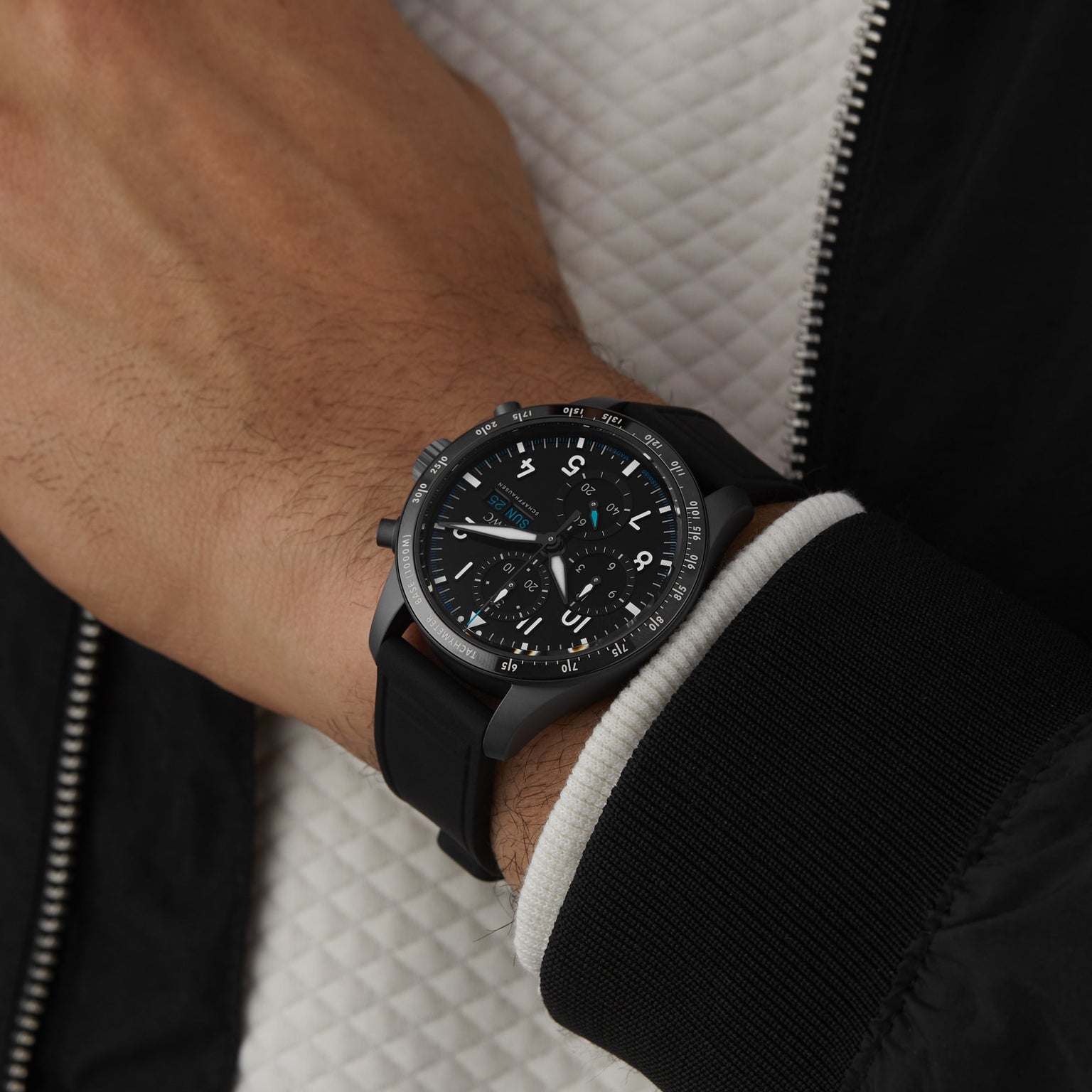 A person wearing the IWC Schaffhausen Pilots Watch Performance Chronograph 41 Mercedes-AMG PETRONAS Formula One™ Team with a black dial and black rubber strap, paired with a black and white jacket.