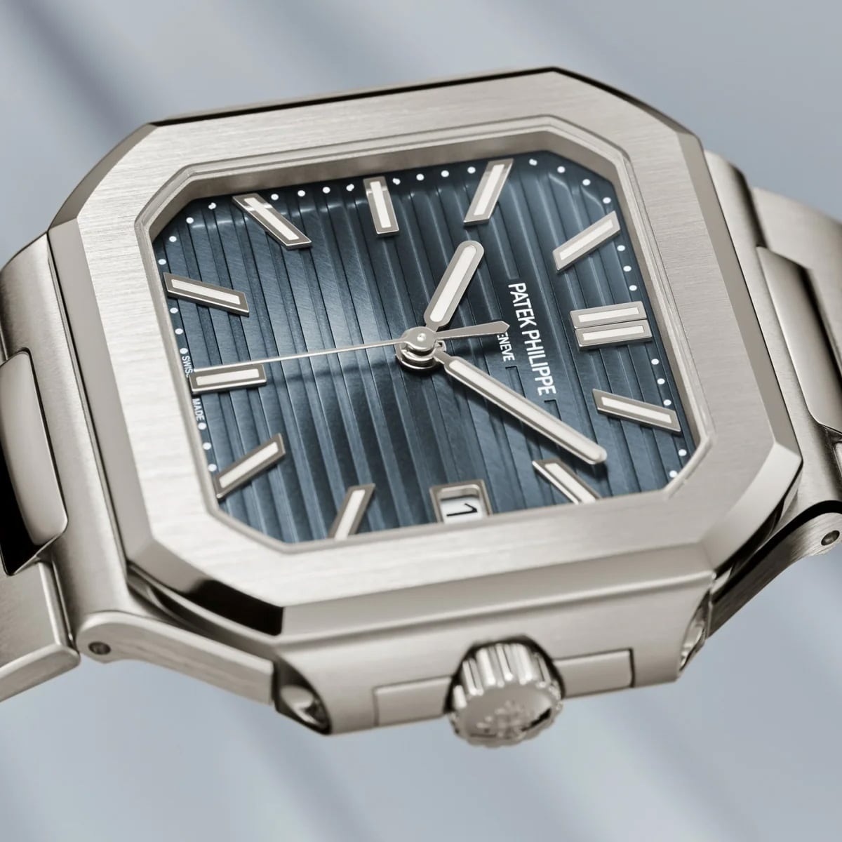 A Patek Philippe Cubitus 7128/1G-001 wristwatch features a silver rectangular case, blue striped dial, and metal bracelet.