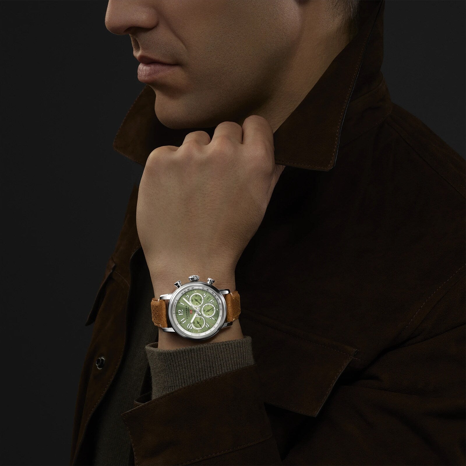 A man wearing a brown jacket displays the Chopard Mille Miglia Classic Chronograph, featuring a green face and tan leather strap.