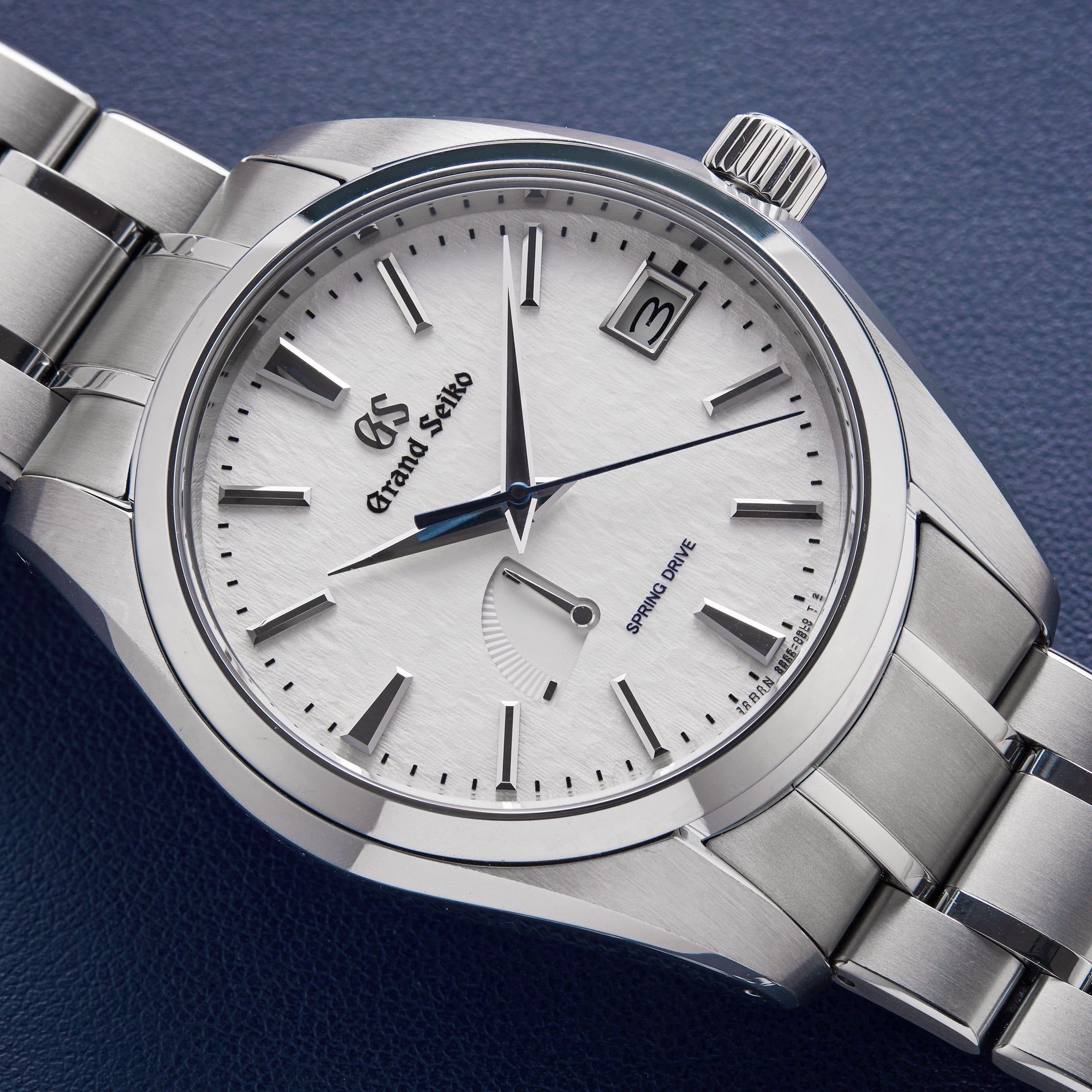 The Grand Seiko Spring Drive Heritage wristwatch featuring a titanium case, silver dial, date at 3 o’clock, power reserve indicator, and a silver metal bracelet rests elegantly on a dark blue surface.