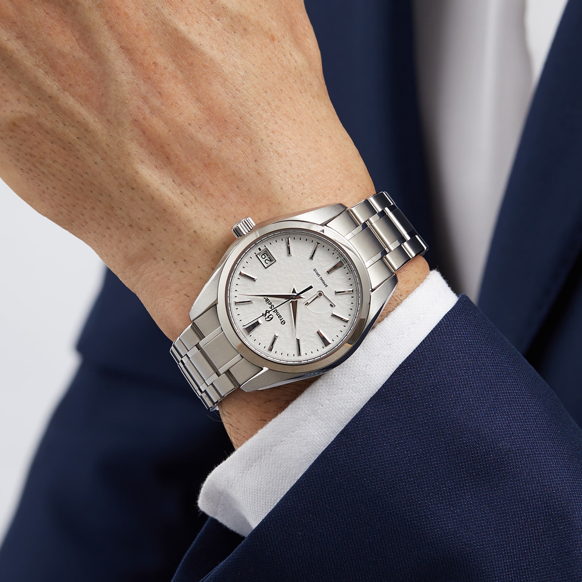 A person wearing a Grand Seiko Spring Drive Heritage watch with a white dial and titanium case, paired with a dark blue suit and white shirt.