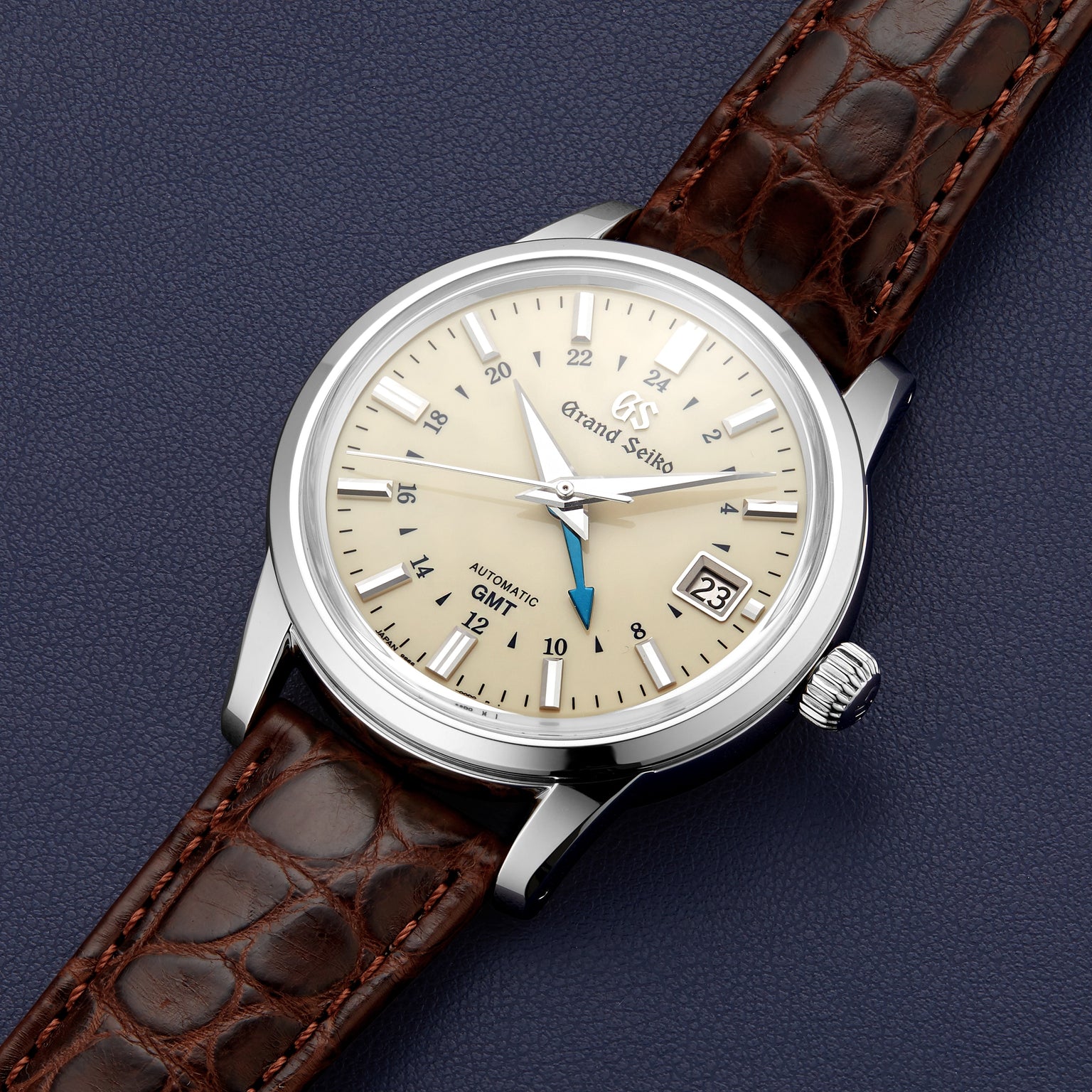 The Grand Seiko Automatic GMT Elegance features a beige dial, brown leather strap, silver case, date at 3 oclock, blue GMT hand, and automatic movement, shown on a dark textured surface.
