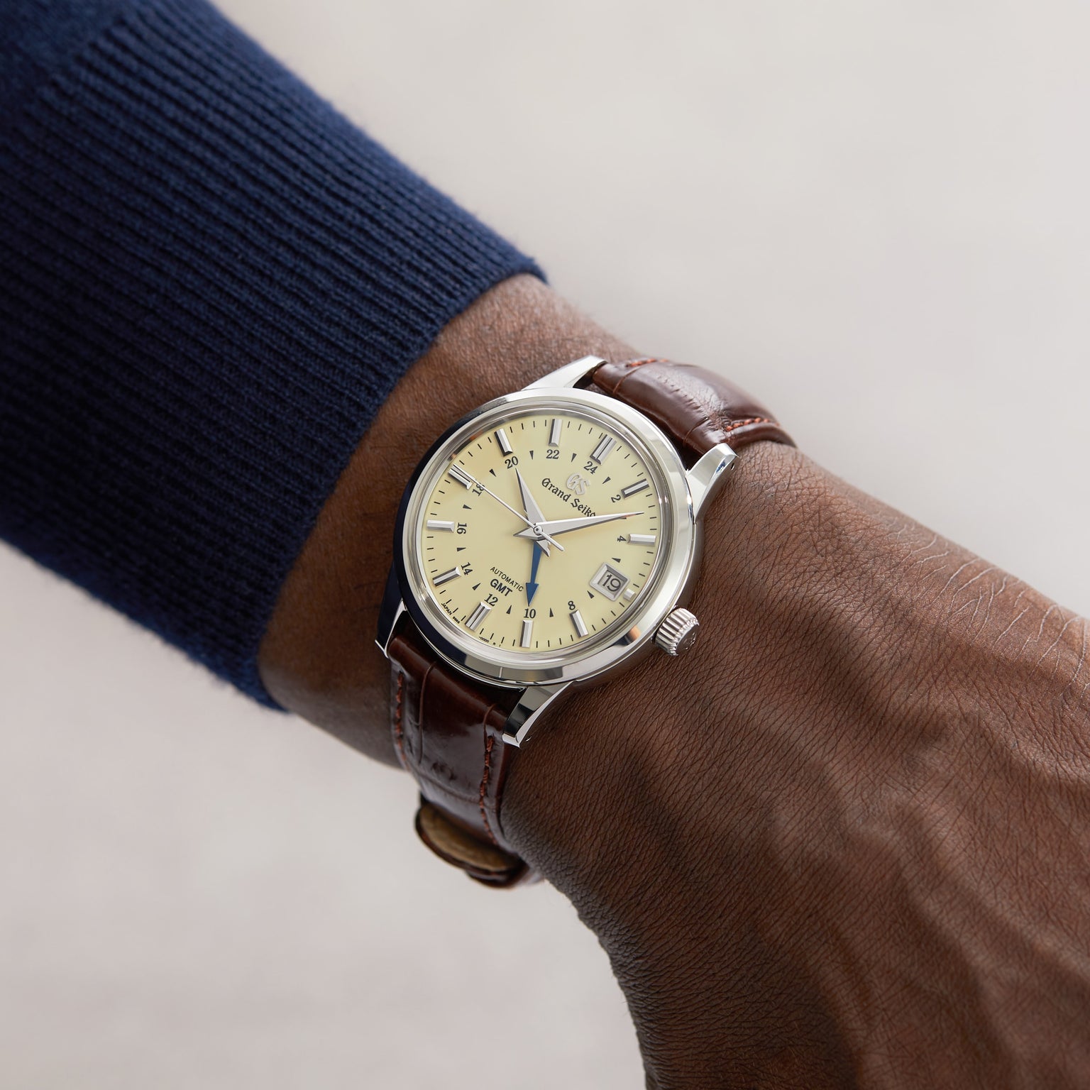 A person wearing a Grand Seiko Automatic GMT Elegance watch from the Elegance Collection, with a beige dial, brown leather strap, automatic movement, and displays for time, day, and date.