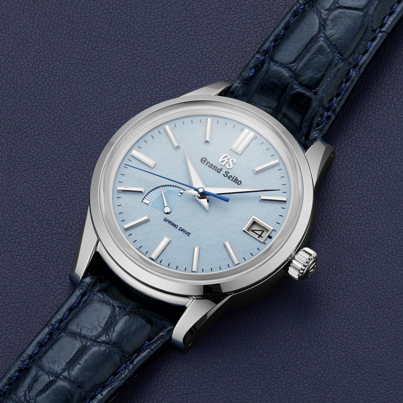 The Grand Seiko Spring Drive Elegance wristwatch features a light blue dial, stainless steel case, silver hands and markers, date at 3 o’clock, and a dark blue leather strap displayed on a textured dark surface.