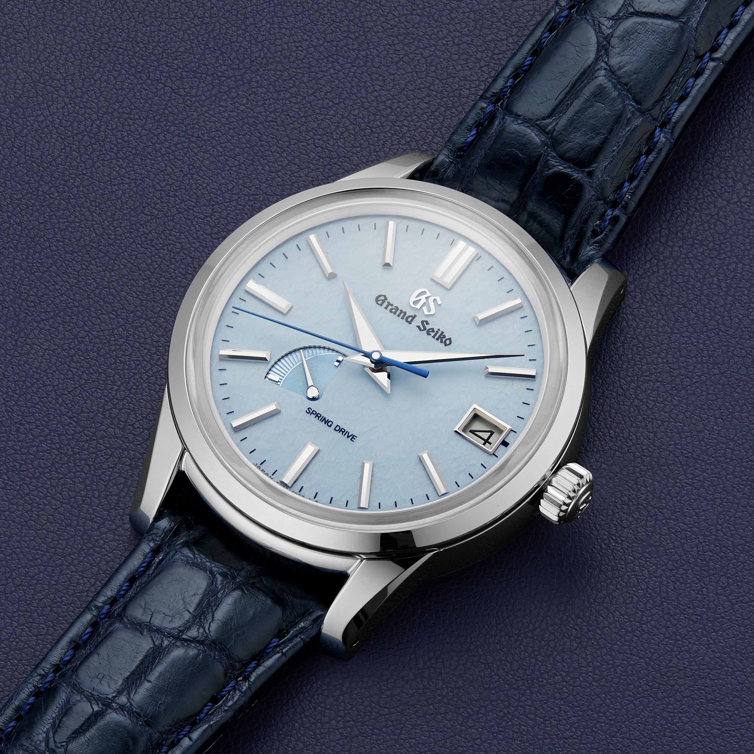 The Grand Seiko Spring Drive Elegance wristwatch features a light blue dial, stainless steel case, silver hands and markers, date at 3 o’clock, and a dark blue leather strap displayed on a textured dark surface.
