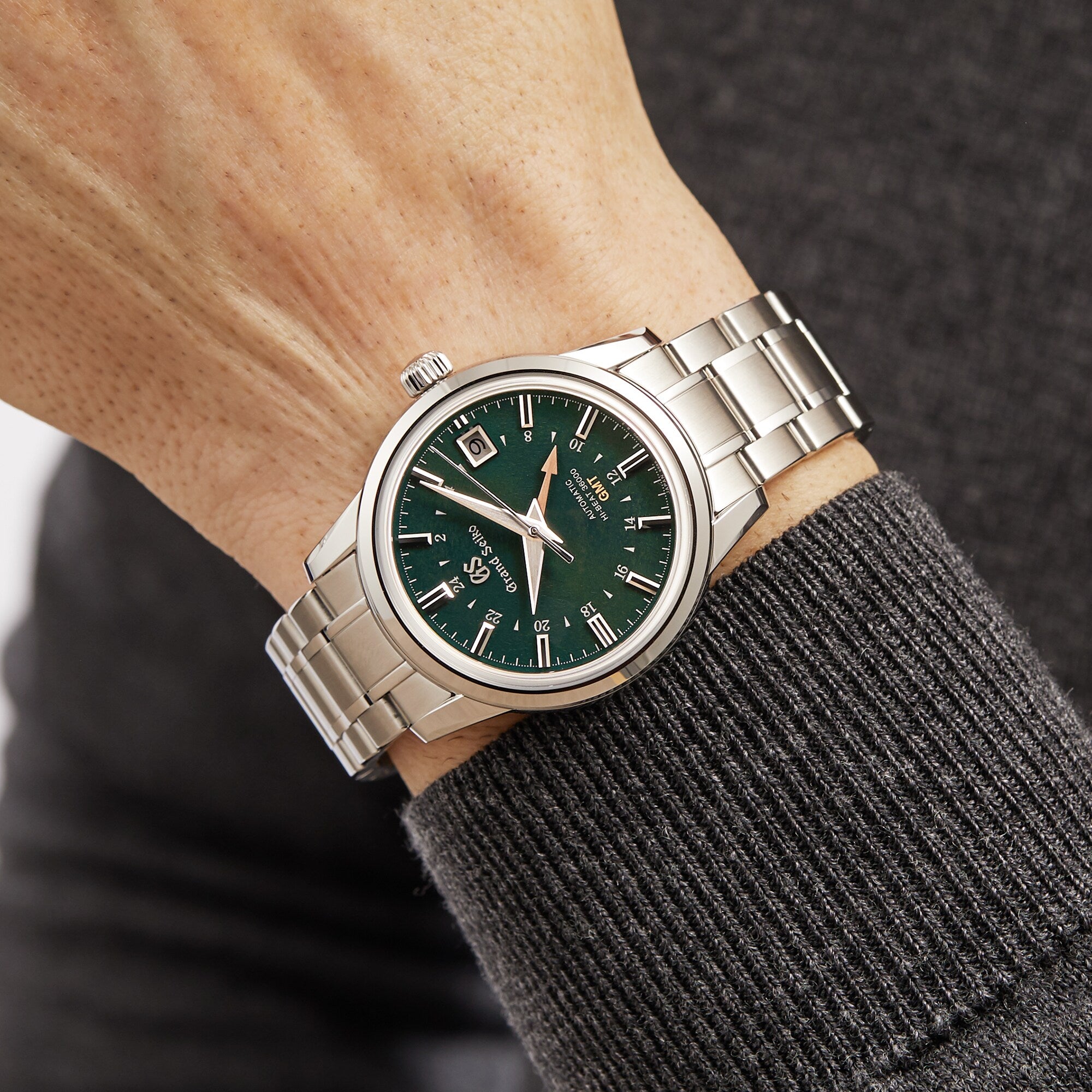 A Grand Seiko Hi-Beat GMT Elegance wristwatch with a green dial and silver hour markers, featuring the Calibre 9S86, is worn on the left wrist against a gray long-sleeve sweater.