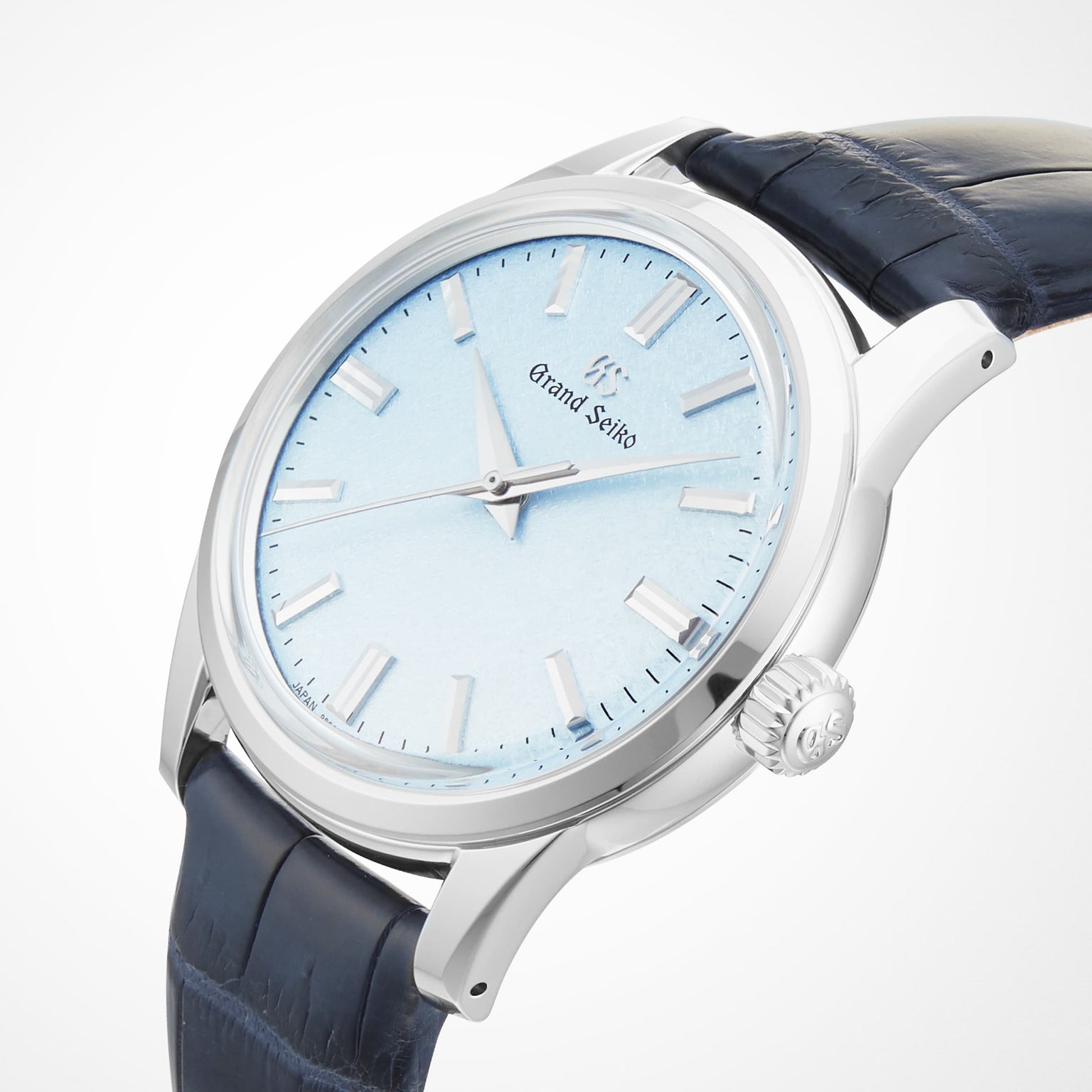 A close-up of the Grand Seiko Manual Elegance watch, featuring a pale blue textured dial, silver hour markers, Caliber 9S64 movement, and a dark blue leather strap.