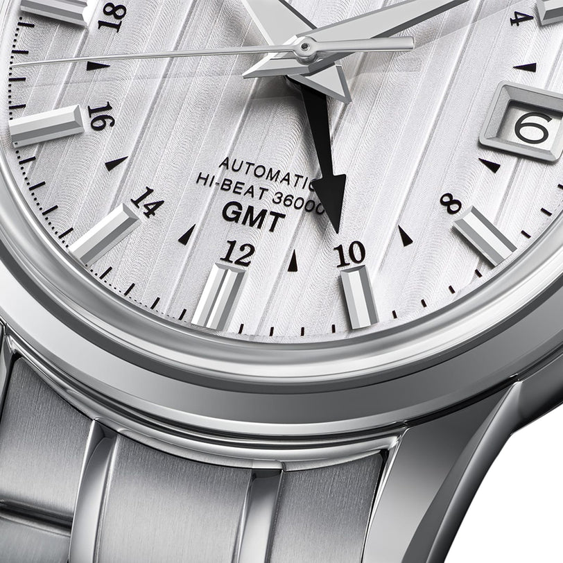 Close-up of the Grand Seiko Hi-Beat GMT Elegance wristwatch, showcasing its textured dial with time, date, and GMT features, plus Automatic Hi-Beat 36000 text and a sleek metal bracelet.