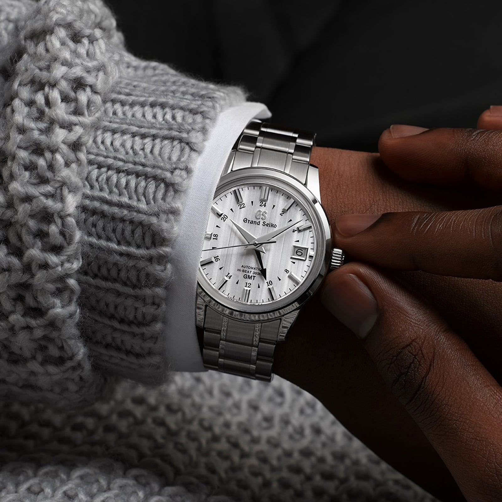 A person wearing a Grand Seiko Hi-Beat GMT Elegance wristwatch with a textured white dial, dressed in a gray knitted sweater and white shirt.