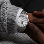 A person wearing a Grand Seiko Hi-Beat GMT Elegance wristwatch with a textured white dial, dressed in a gray knitted sweater and white shirt.