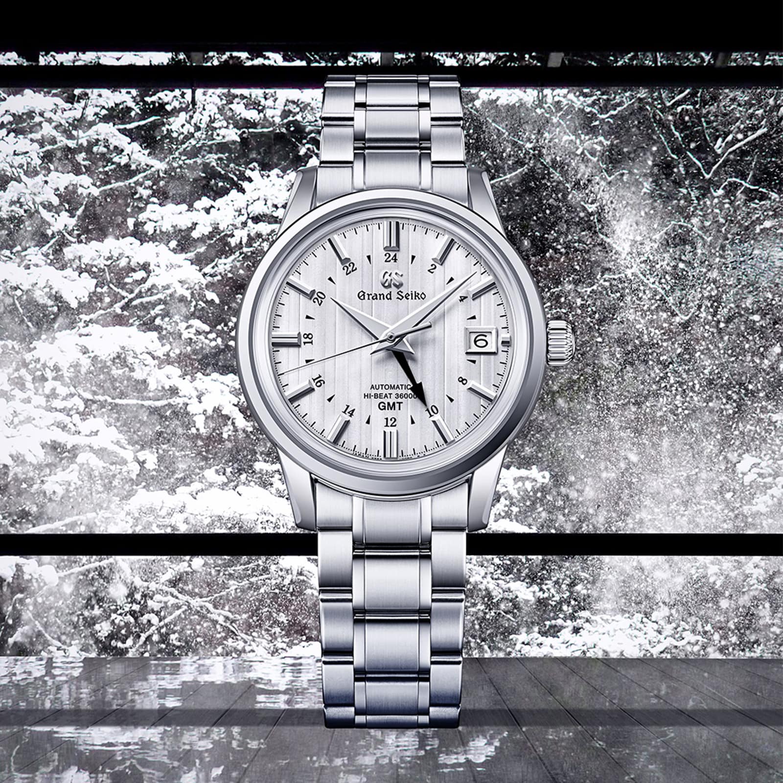 A Grand Seiko Hi-Beat GMT Elegance stainless steel wristwatch with a white textured dial, shown against a snowy window backdrop.