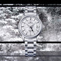 A Grand Seiko Hi-Beat GMT Elegance stainless steel wristwatch with a white textured dial, shown against a snowy window backdrop.