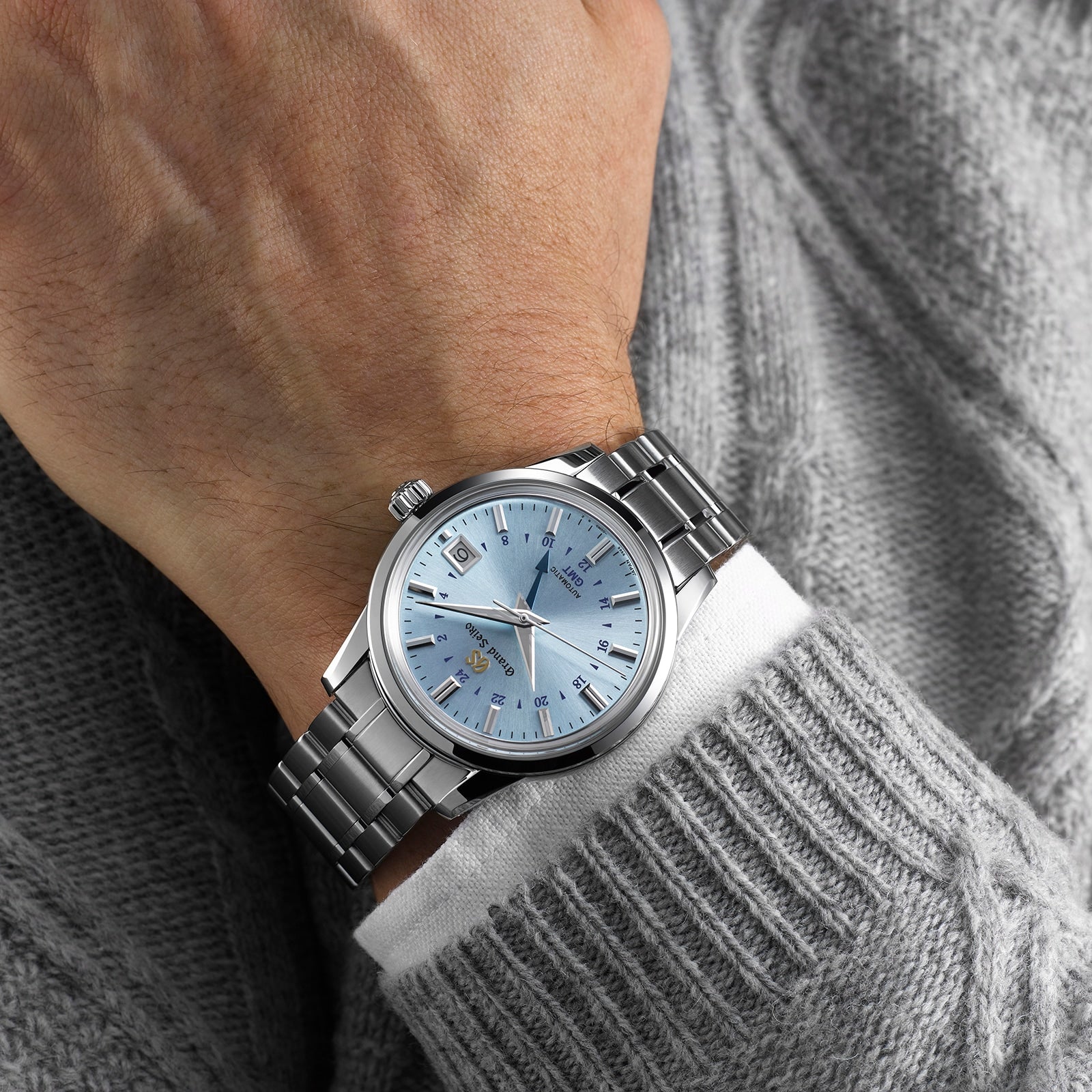 A person wearing the Grand Seiko Automatic GMT Elegance with a light blue dial and silver bracelet, styled over the sleeve of a grey textured sweater.