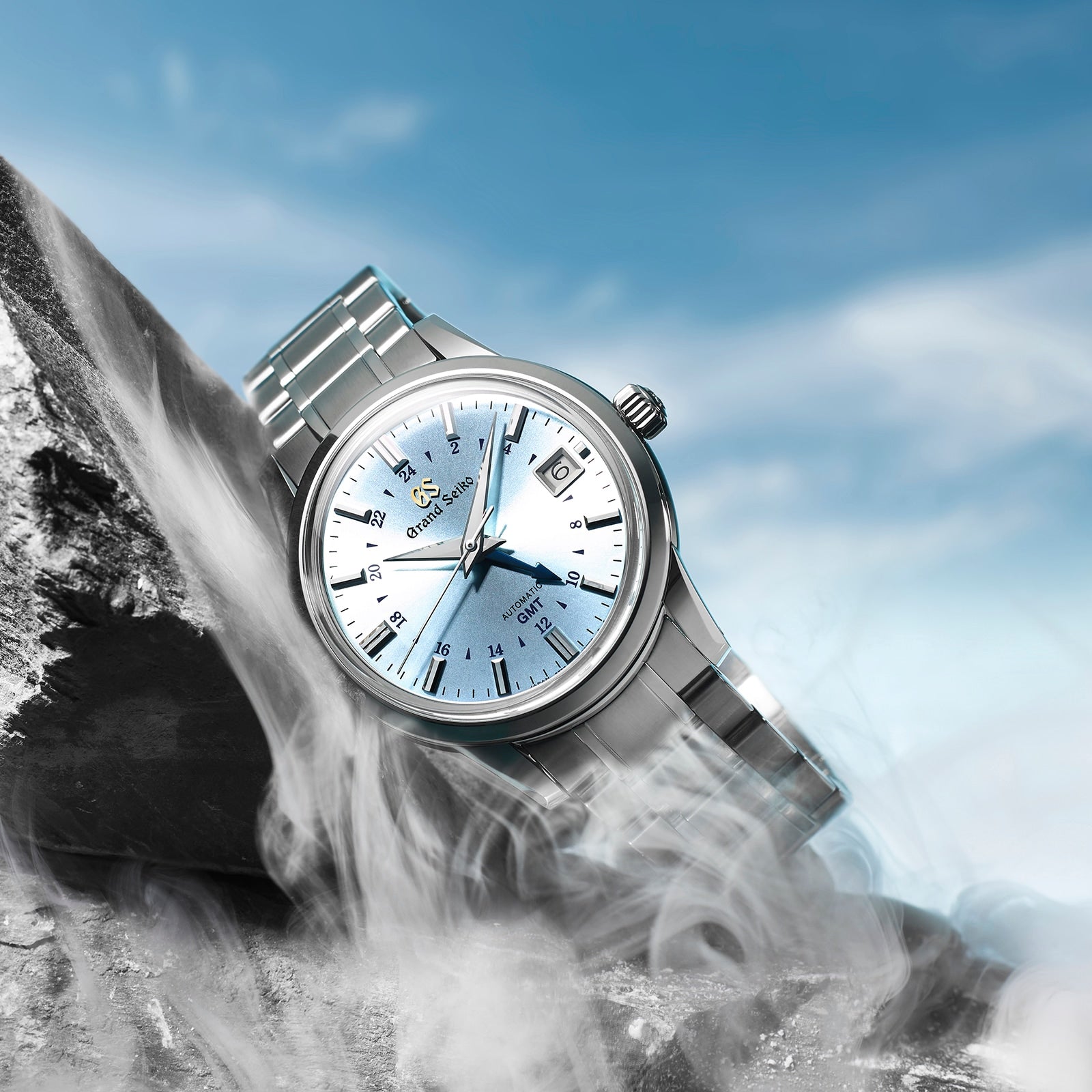 The Grand Seiko Automatic GMT Elegance, featuring a light blue face, rests on a rock with swirling mist and a blue sky behind—a refined timepiece crafted for sophistication.