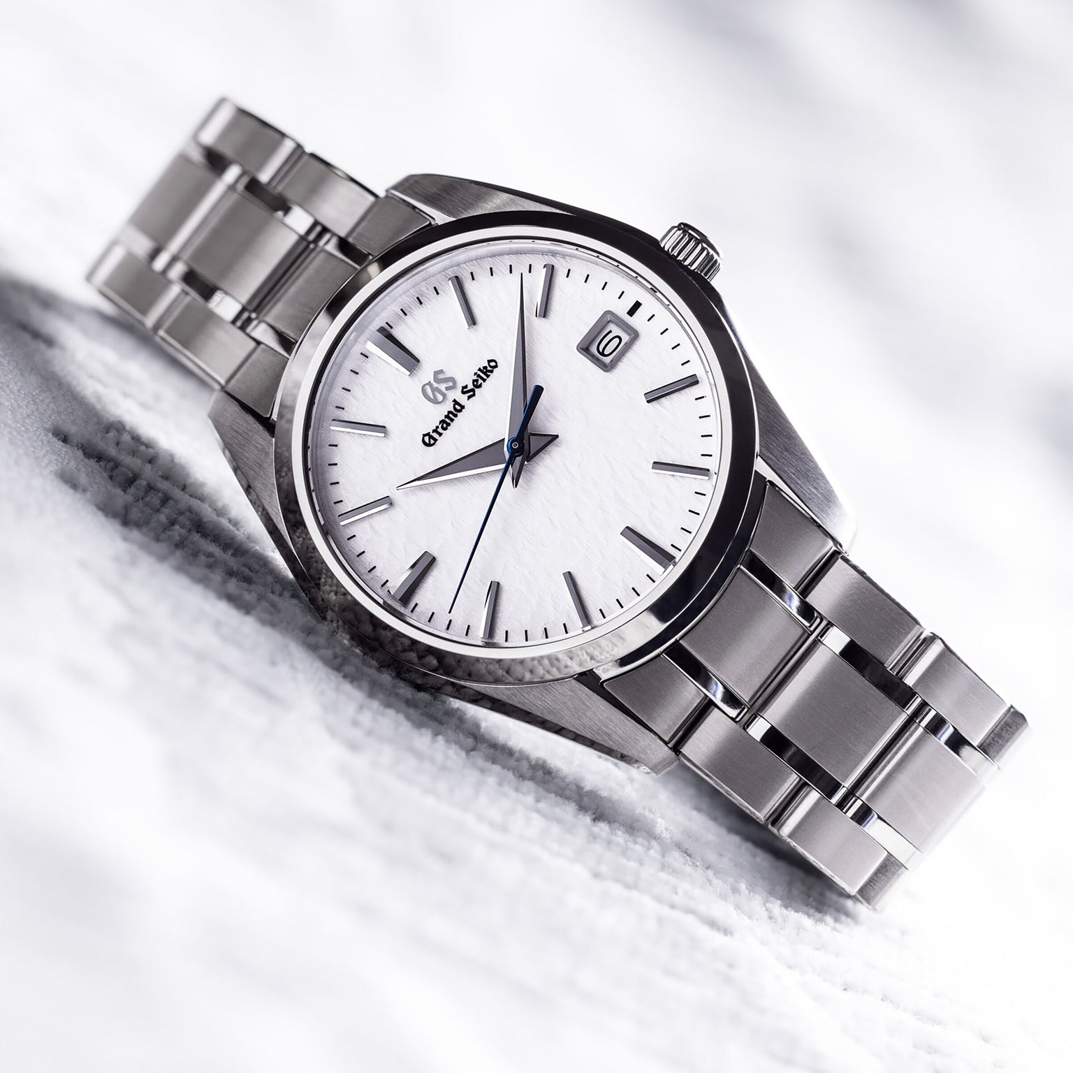 The Grand Seiko Quartz Heritage wristwatch showcases a 37mm titanium case, white snowflake dial, silver hour markers, and a date window at 3 o’clock, displayed on a light surface.