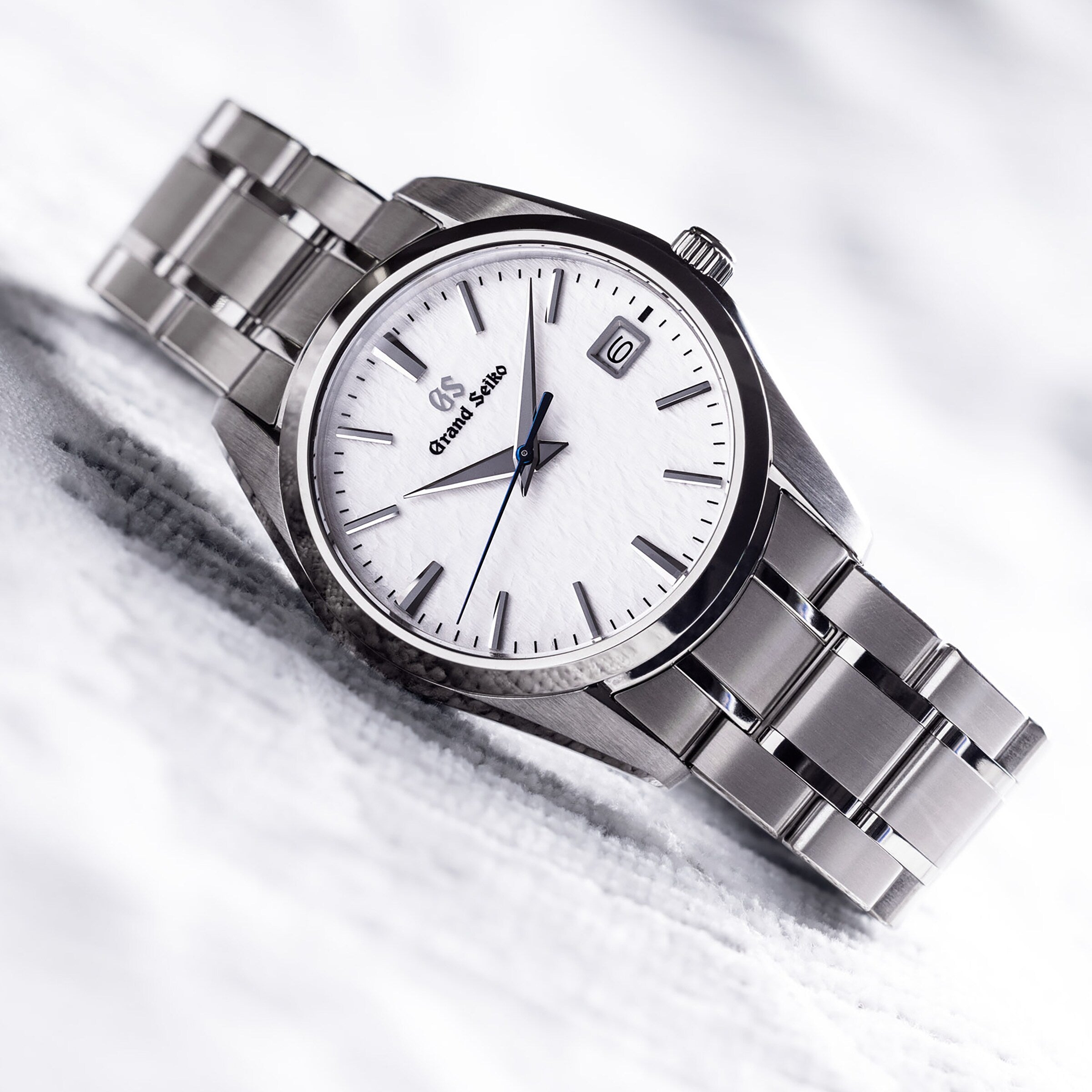 The Grand Seiko Quartz Heritage wristwatch showcases a 37mm titanium case, white snowflake dial, silver hour markers, and a date window at 3 o’clock, displayed on a light surface.