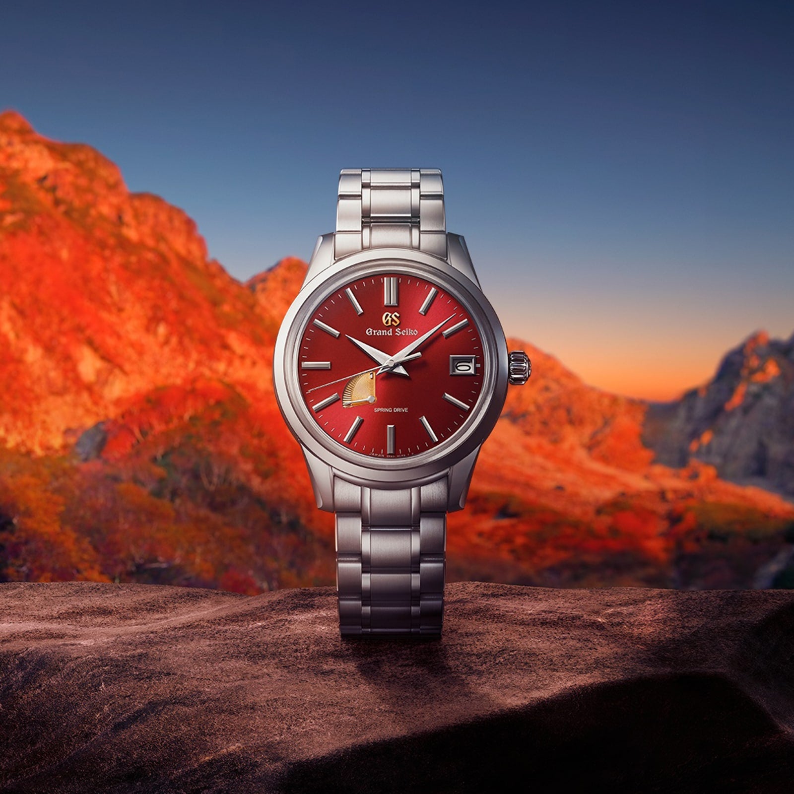 The Grand Seiko Spring Drive Elegance, featuring a silver case and red dial, is showcased before a vivid sunset over mountain peaks.