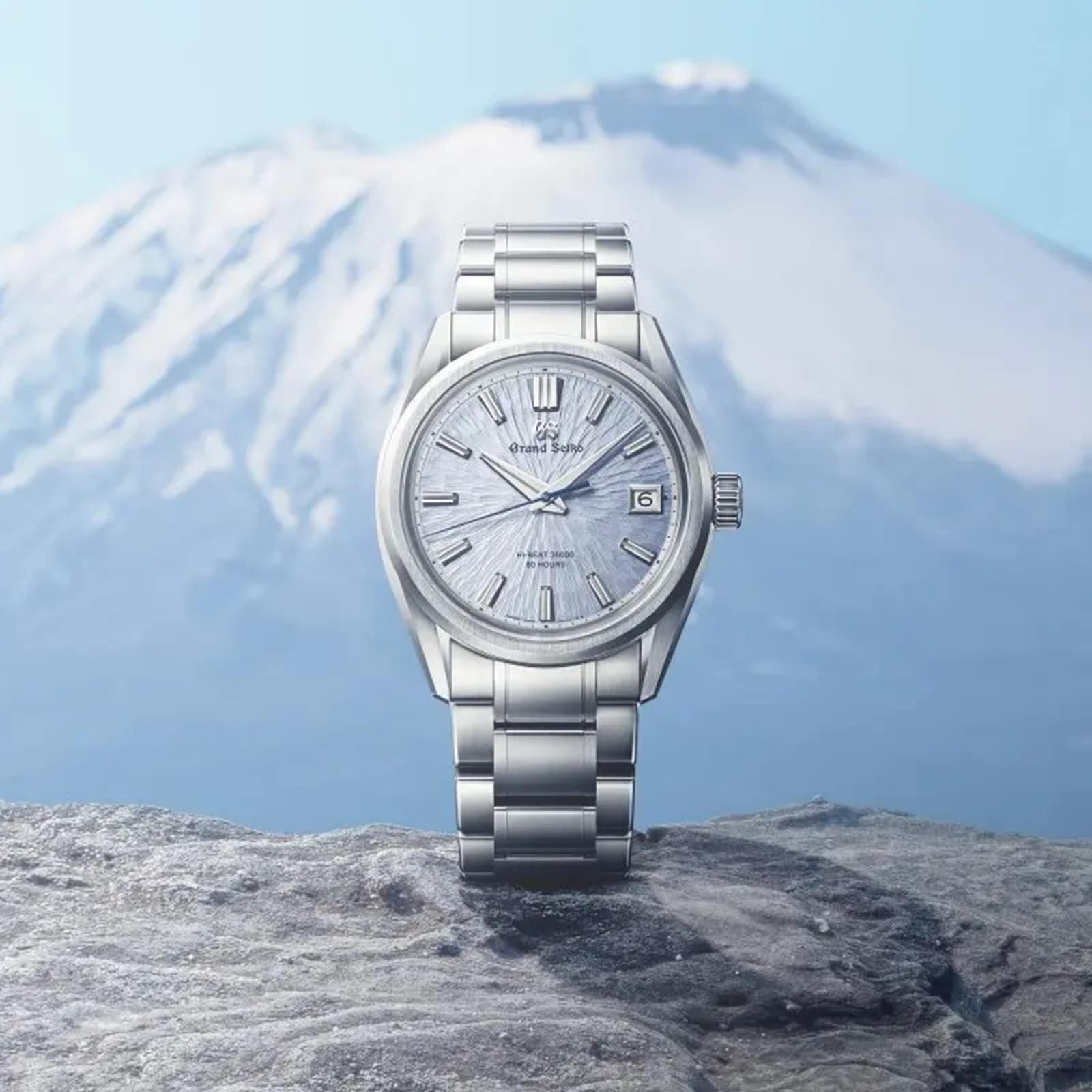 The Grand Seiko Hi-Beat 80 Hours Evolution 9 wristwatch with a textured silver face is displayed on a rock, set before the blurred backdrop of a snow-capped mountain landscape.