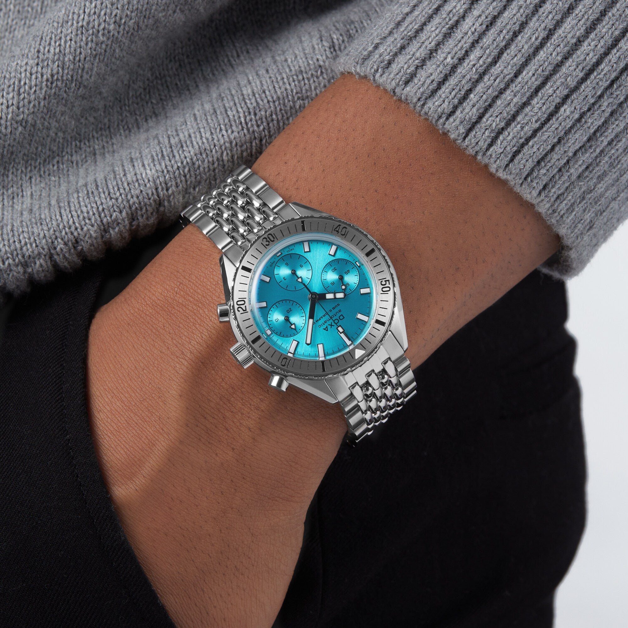 A person wearing the DOXA Sub 200 C-Graph II Aquamarine, a mechanical automatic chronograph with a turquoise dial and stainless steel bracelet, styled with a grey sweater and black pants.