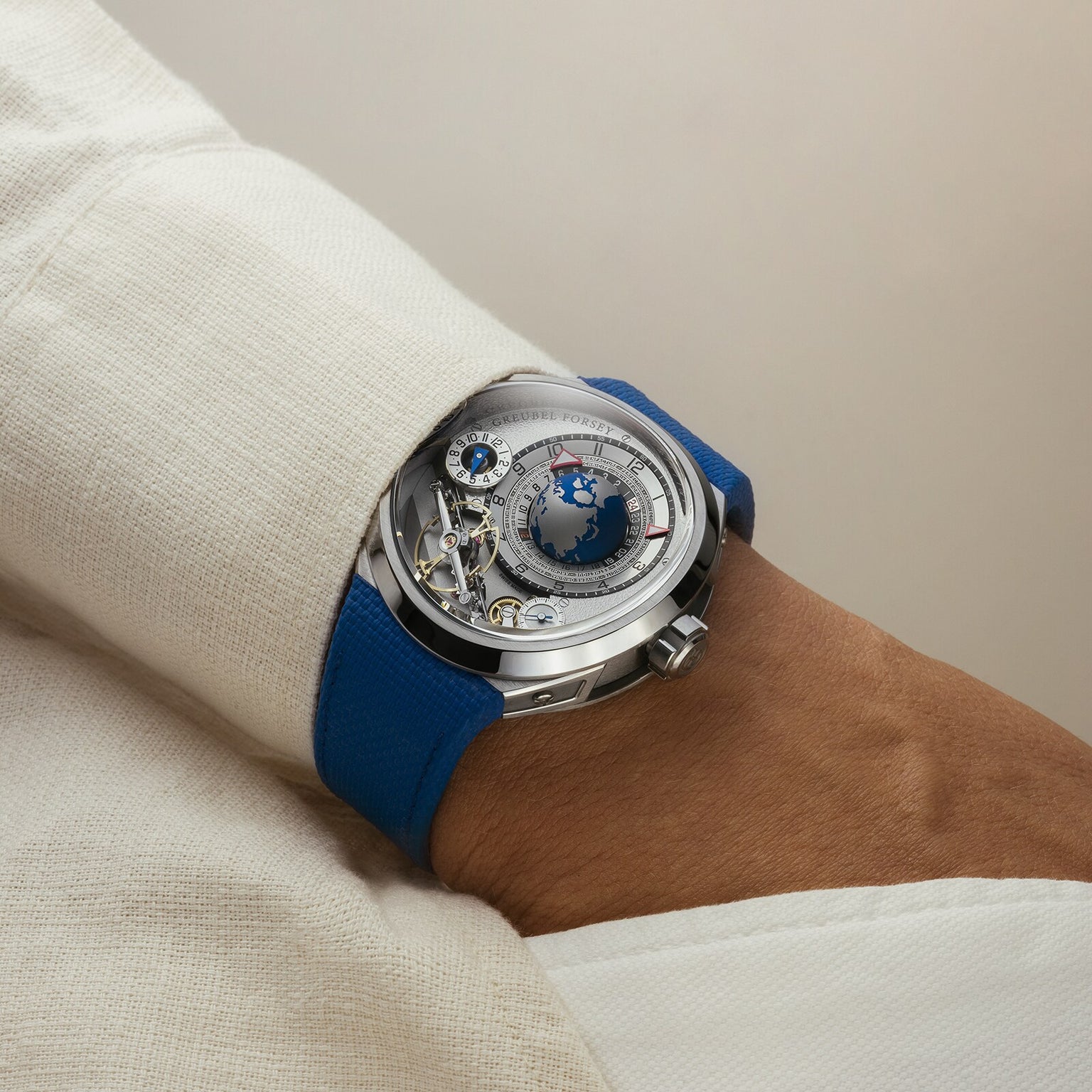 The Greubel Forsey GMT Balancier Convexe, a silver luxury watch with a blue strap, is worn on a wrist in a cream-colored sleeve.