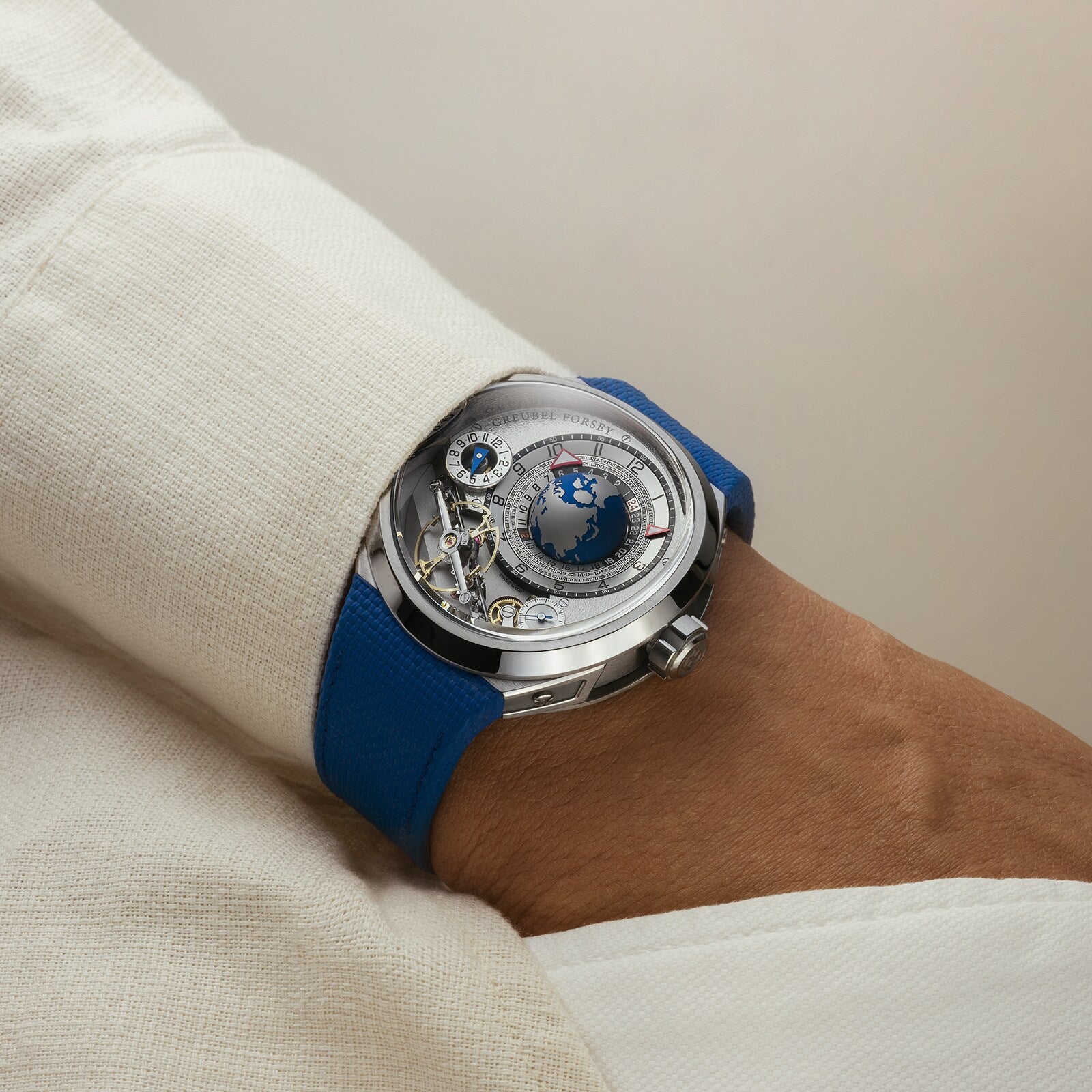 The Greubel Forsey GMT Balancier Convexe, a silver luxury watch with a blue strap, is worn on a wrist in a cream-colored sleeve.