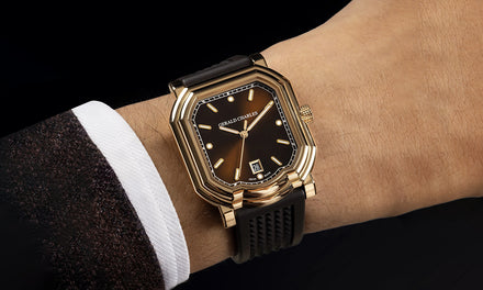 A wrist with a brown coat and white shirt sleeve wears a Gerald Charles Maestro 2.0 watch with brown strap and gold bezel and brown metallic dial