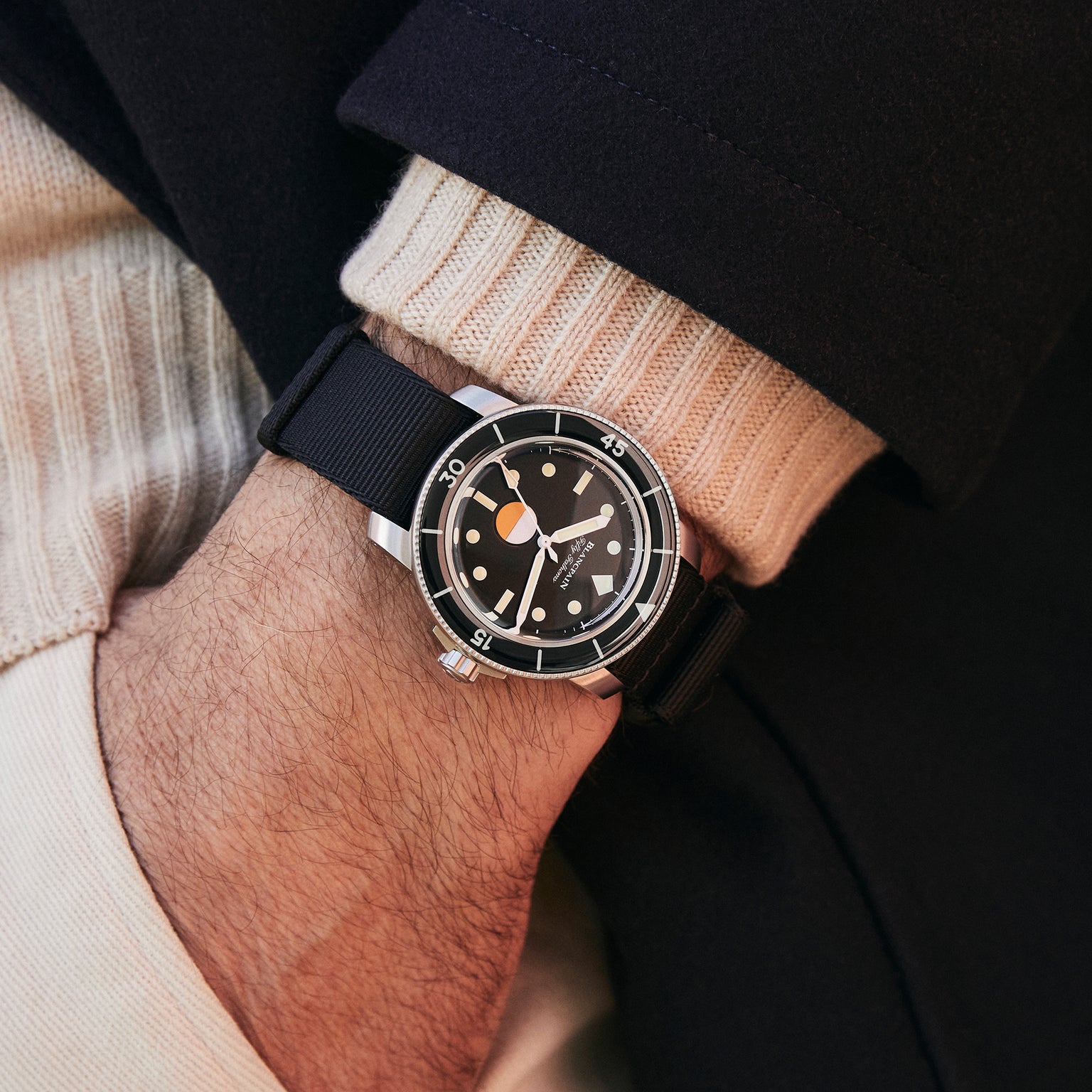 A person wearing a Blancpain Fifty Fathoms MIL-SPEC Limited Edition For HODINKEE watch with a black strap, paired with a cream sweater and dark coat.
