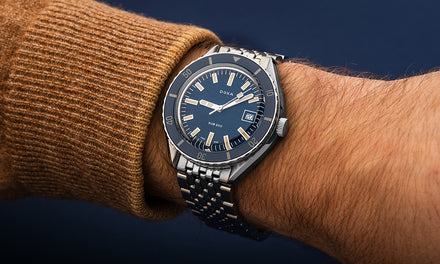 A DOXA Sub 200 watch on a person's wrist with a brown sweater over a blue background