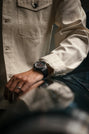 A person in a cream shirt wears the Porsche Design Chronograph 1 Hodinkee 2024 Edition on their wrist, resting their hand on a shiny surface.