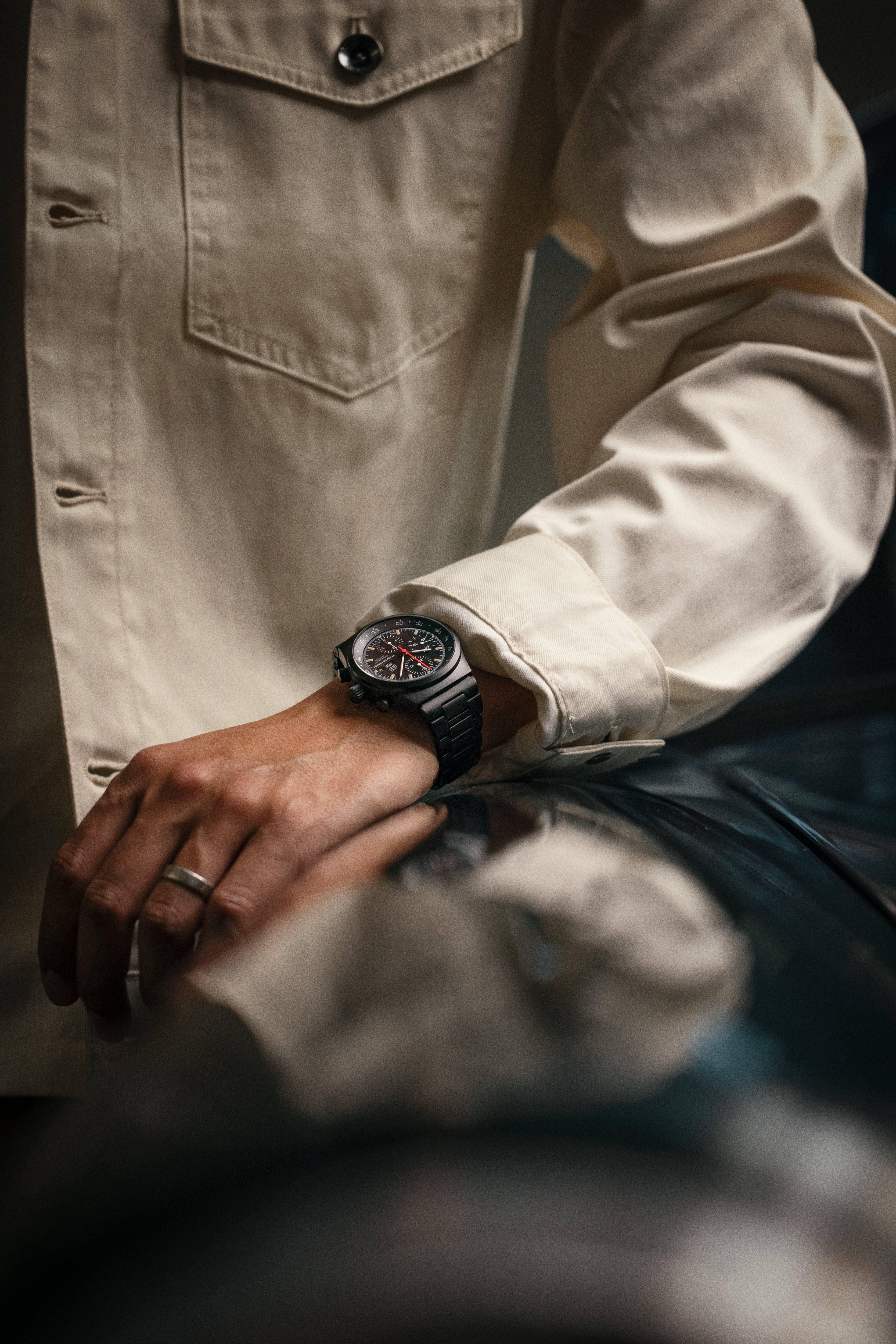 A person in a cream shirt wears the Porsche Design Chronograph 1 Hodinkee 2024 Edition on their wrist, resting their hand on a shiny surface.