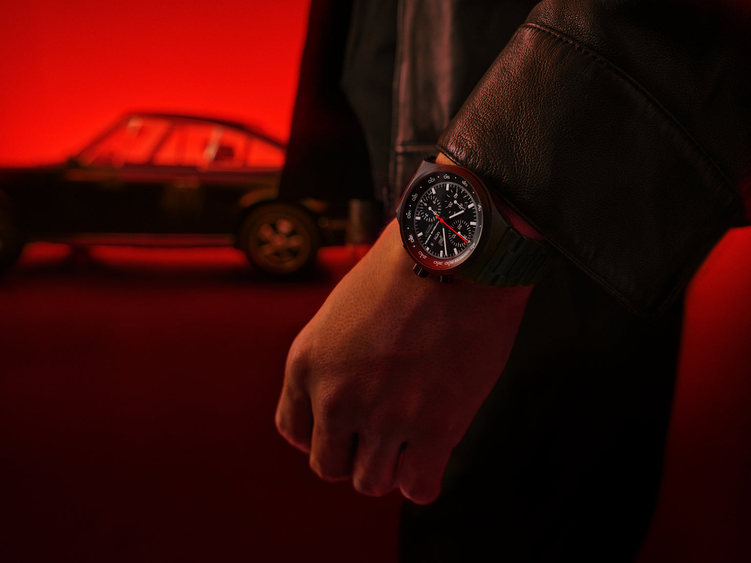 A person wearing the Porsche Design Chronograph 1 All Black Numbered Edition stands in front of a blurred car with a red background.