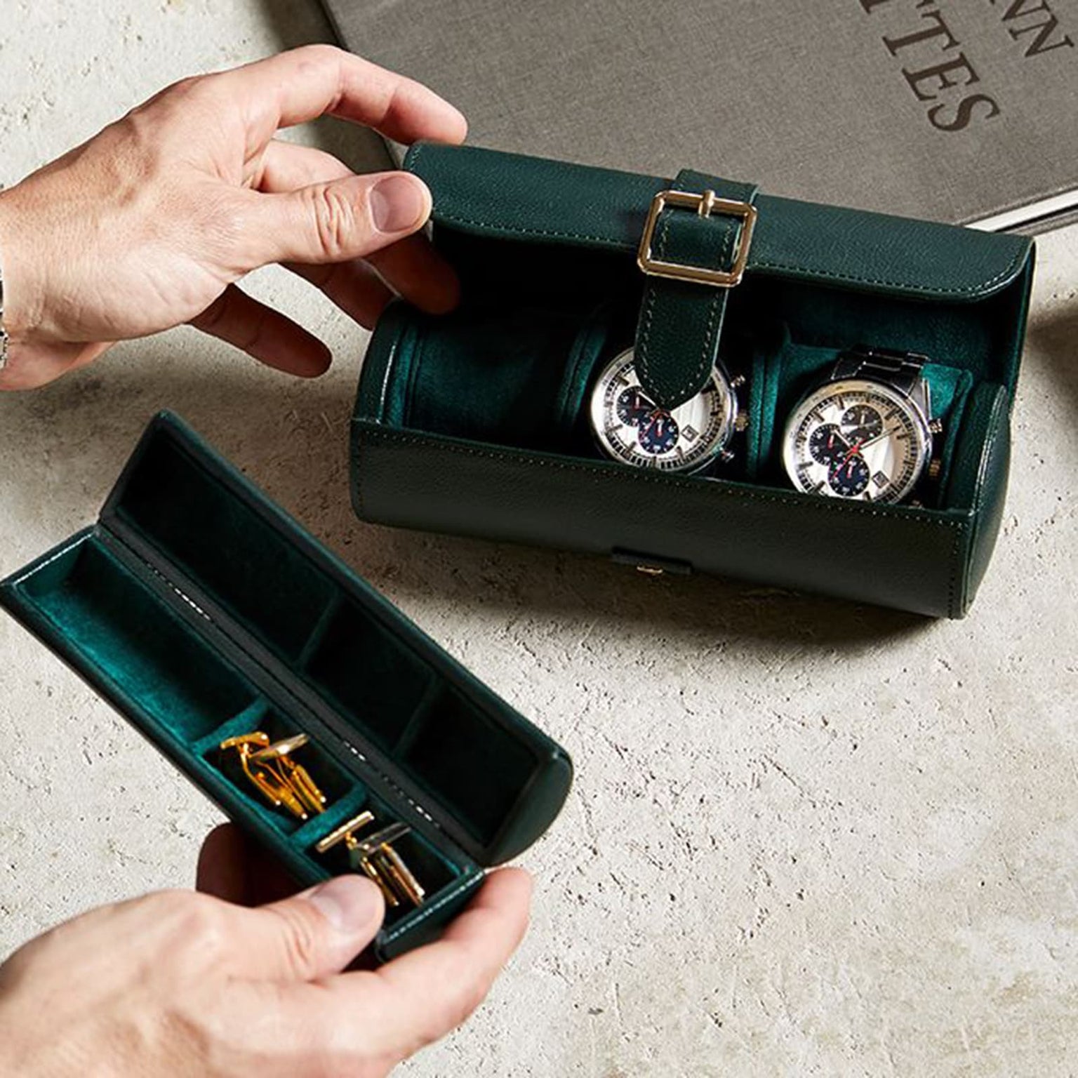 Hands holding the WOLF British Racing Triple Watch Roll in green leather with two watches, alongside another case containing gold cufflinks, all placed on a stone surface.