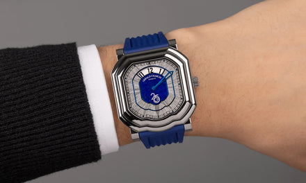A wrist with a white shirt sleeve and a black sweater wears the Gerald Charles Maestro 4.0 Anniversary edition watch with a blue strap and silver toned bezel and blue and gray dial