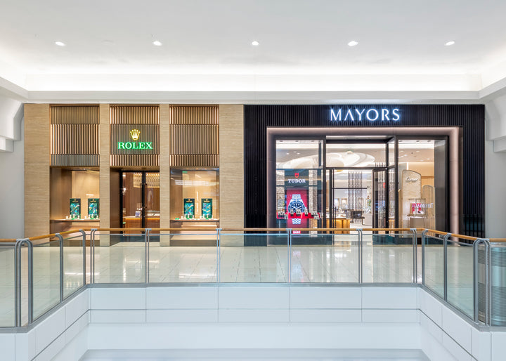 Storefronts of Rolex and Mayors jewelry stores in a shopping mall, with glass displays and bright lighting.