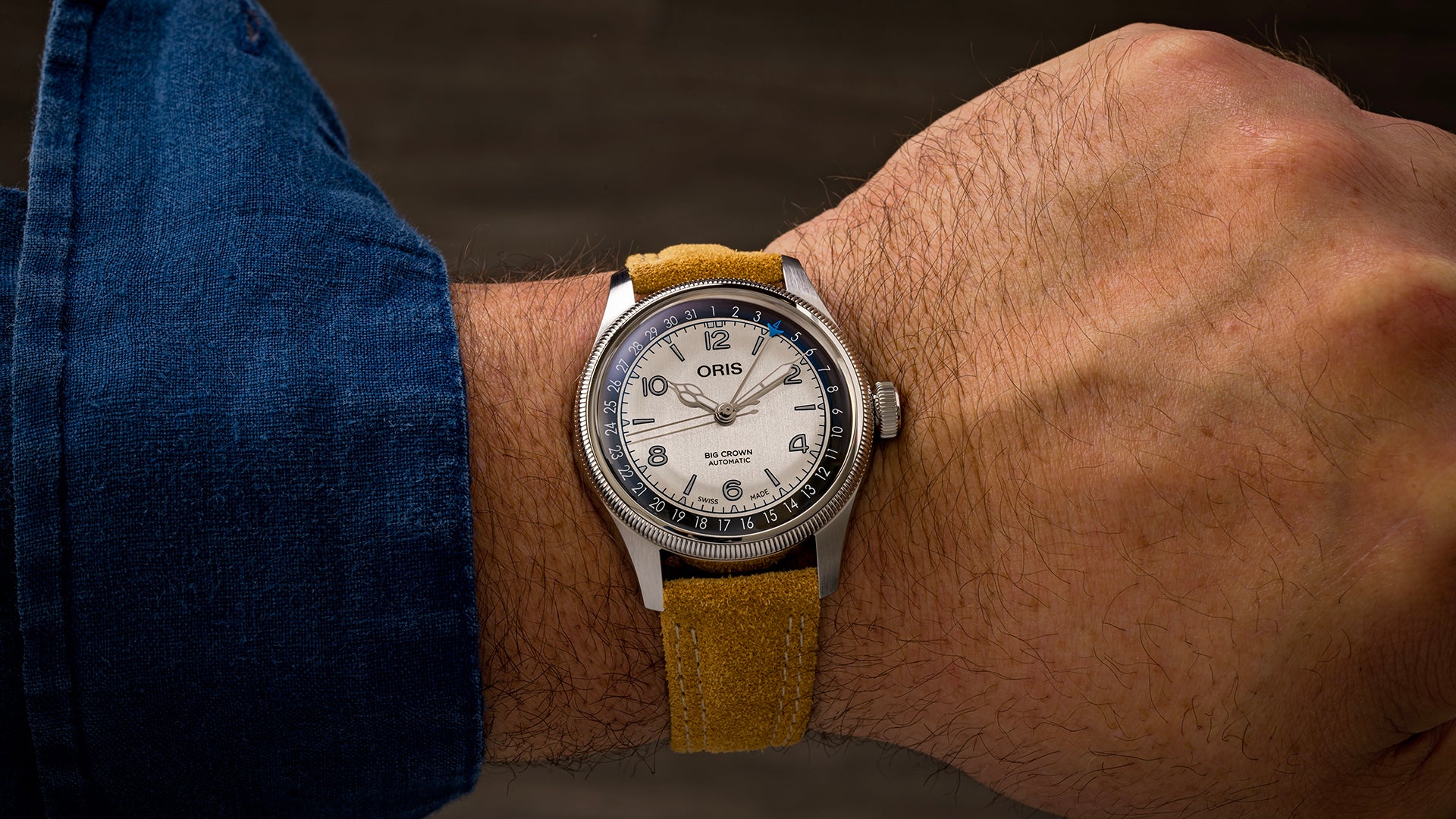 Oris watch with white dial, Arabic numerals, and tan suede strap on wrist wearing blue denim sleeve.