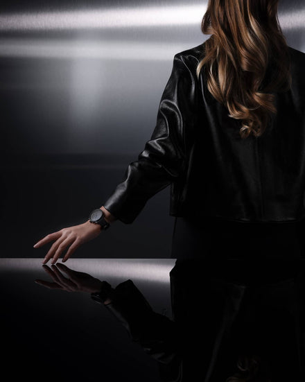 Person wearing a black shirt and watch, standing in a dark room with a reflective surface.