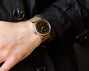 Close-up of a person wearing a watch with a dark face and brown leather strap, against a dark background.