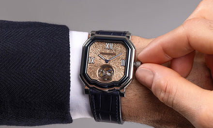 A person's hand adjusts the crown of a Gerald Charles watch with a black strap and textured gold dial with an exposed tourbillon mechanism