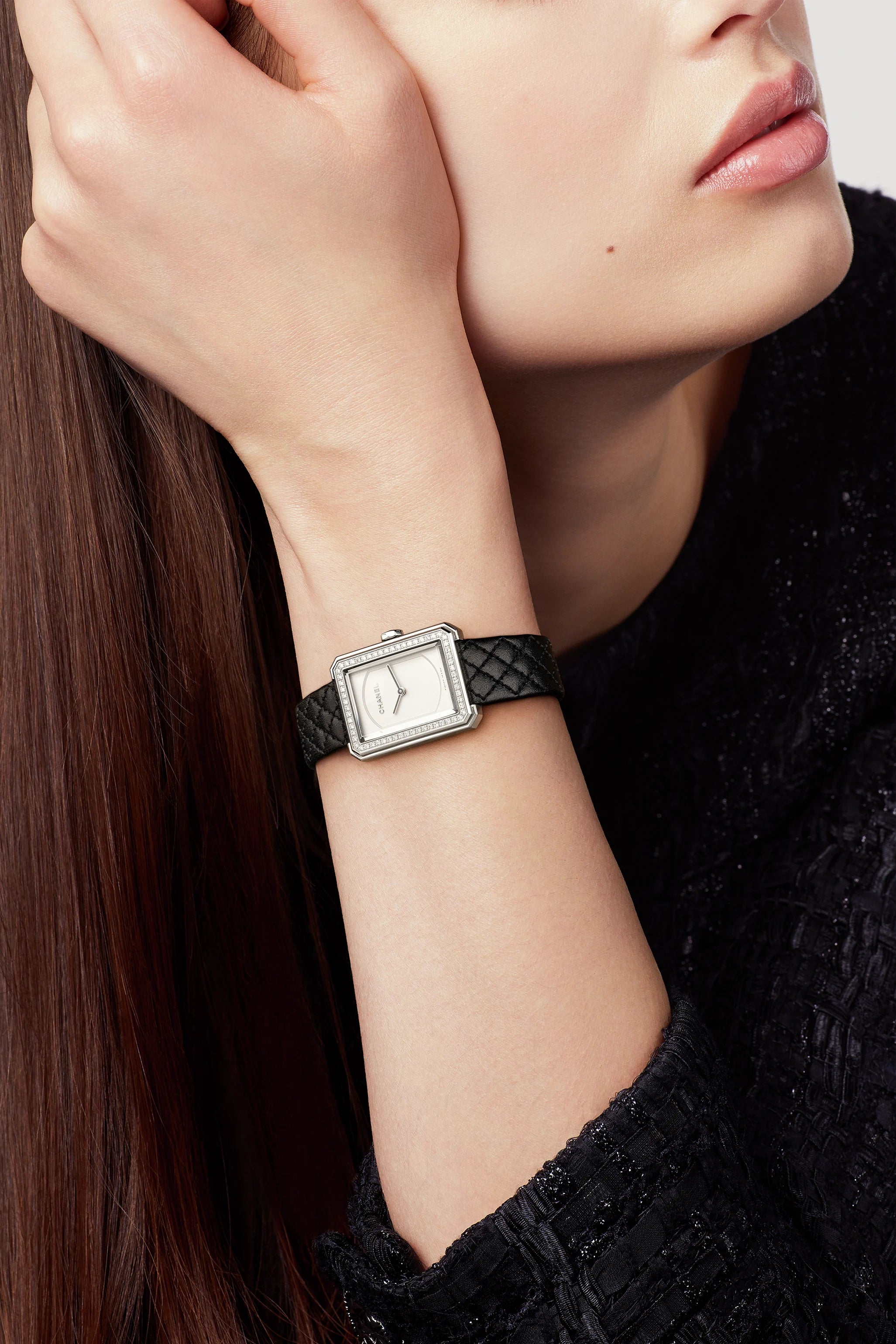 A woman with straight brown hair, wearing a textured black top, rests her chin on her hand and shows off the Chanel Boy·Friend Small Model watch with a square face and black calfskin strap. She has a neutral expression.