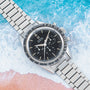 An OMEGA Speedmaster stainless steel watch with a black dial set against a beach-themed background.