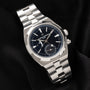 A Vacheron Constantin Overseas Dual-Time wristwatch in silver with a black dial and date display, shown on black fabric.