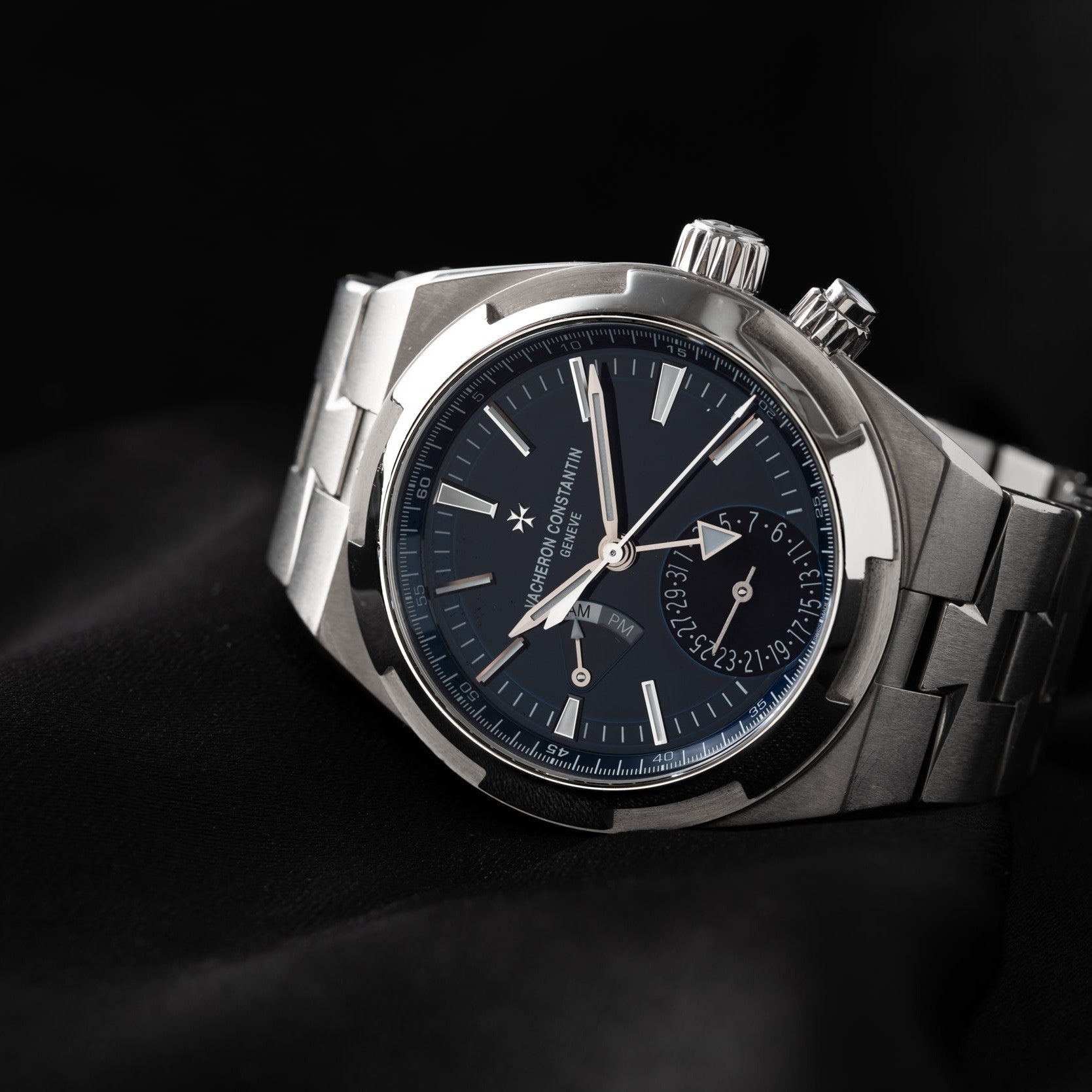 The Vacheron Constantin Overseas Dual-Time, a luxury silver wristwatch with a blue dial and metal band, is showcased on a black fabric background.