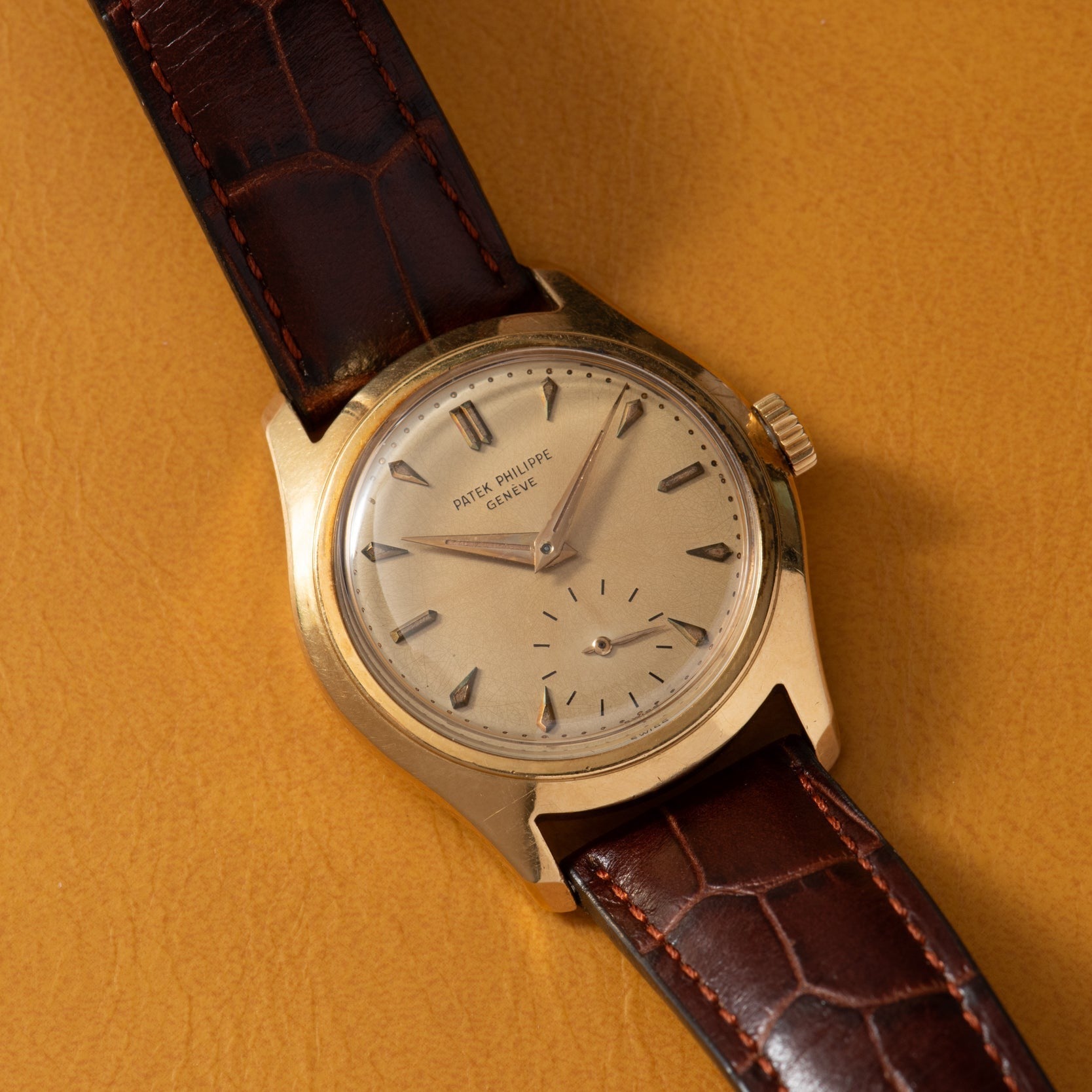 A Patek Philippe Calatrava gold wristwatch with a brown leather strap rests on a tan surface.