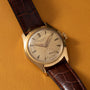 A Patek Philippe Calatrava gold wristwatch with a brown leather strap rests on a tan surface.
