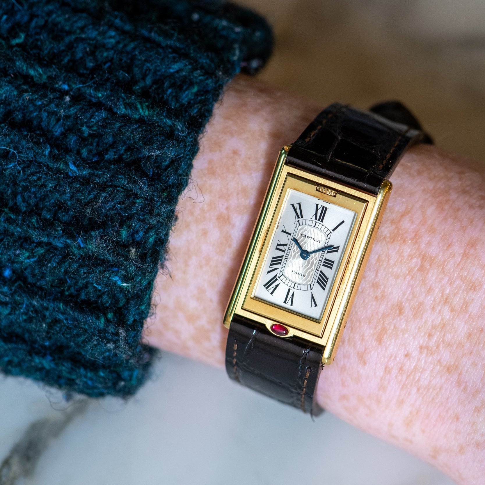 The Cartier Tank Basculante Mécanique 150th Anniversary Limited Edition is a gold rectangular wristwatch with Roman numerals on a brown leather strap, shown worn on a person’s wrist.
